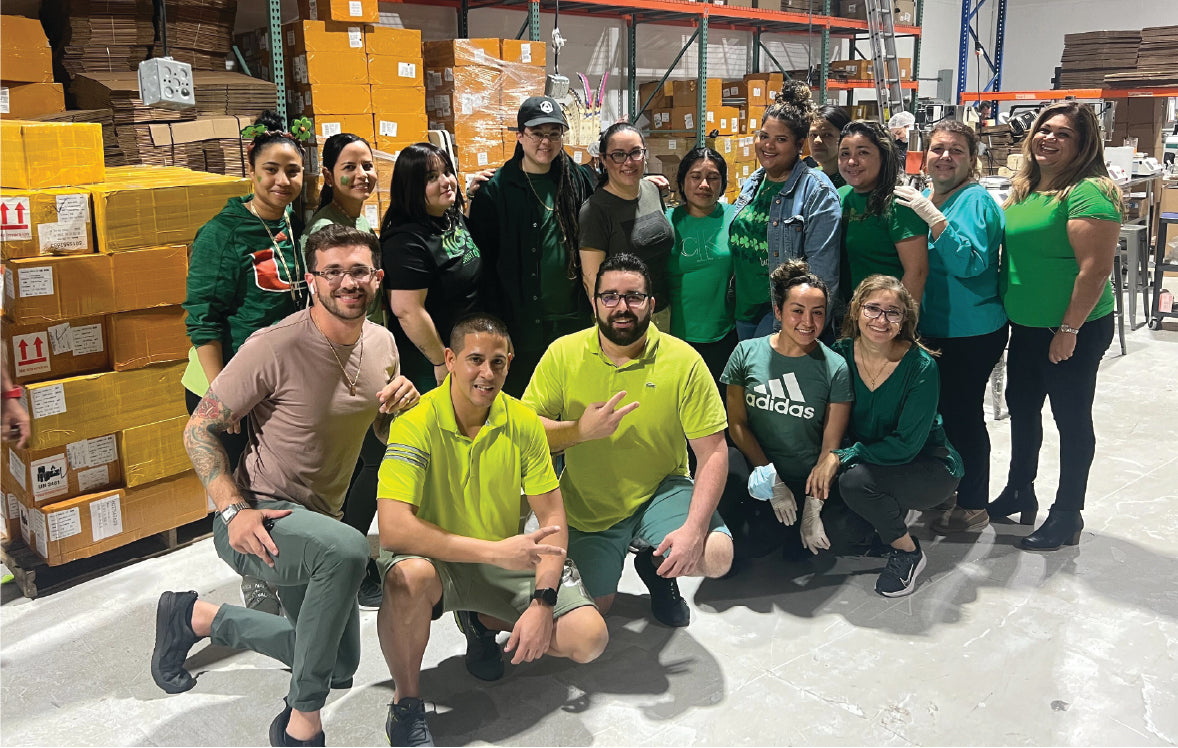 A group of people wearing green and yellow shirts pose together in a warehouse-like environment. They are surrounded by stacked orange boxes and industrial shelves. Most are smiling, appearing cheerful and friendly.