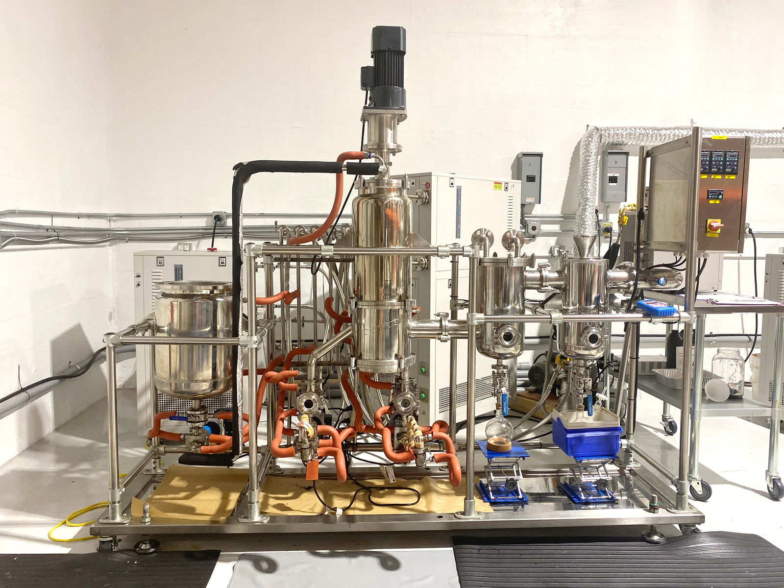 A complex industrial machine with stainless steel components and exposed orange wiring is set up against a white wall. Multiple containers, valves, and control boxes are visible, suggesting a processing or manufacturing setup.
