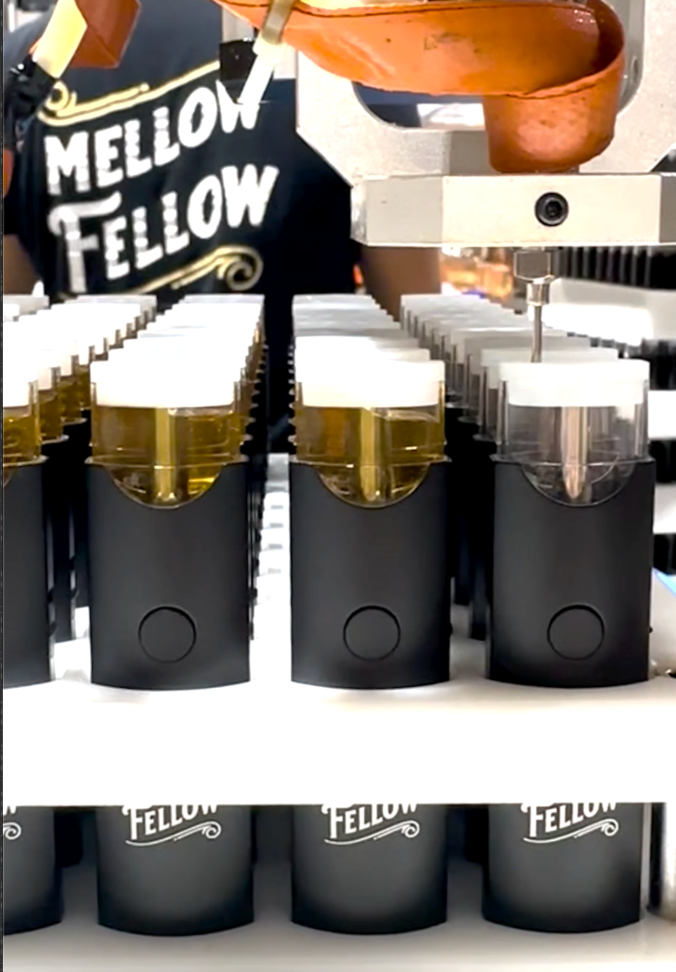 A machine is filling vape cartridges with liquid. The cartridges are lined up in rows, and a person wearing a "Mellow Fellow" shirt is operating the machine in the background.