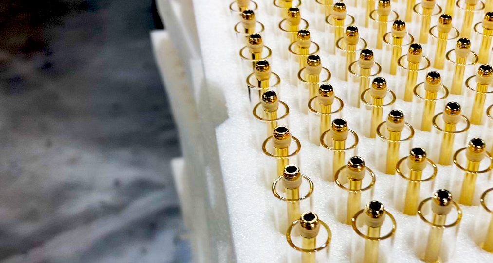 Rows of golden pins with black tips are neatly arranged in a white foam holder. The background is a blurred gray surface, highlighting the organized and reflective nature of the pins' arrangement.