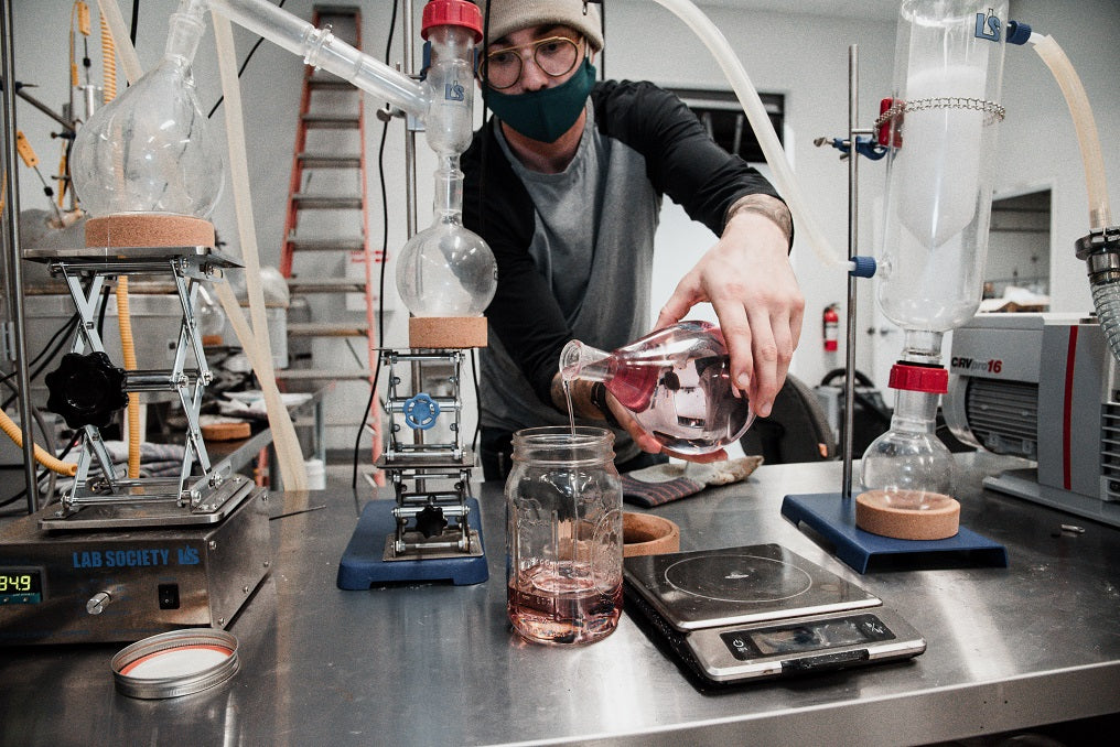 Person in a lab, wearing a mask and beanie, pouring edible oil into a jar