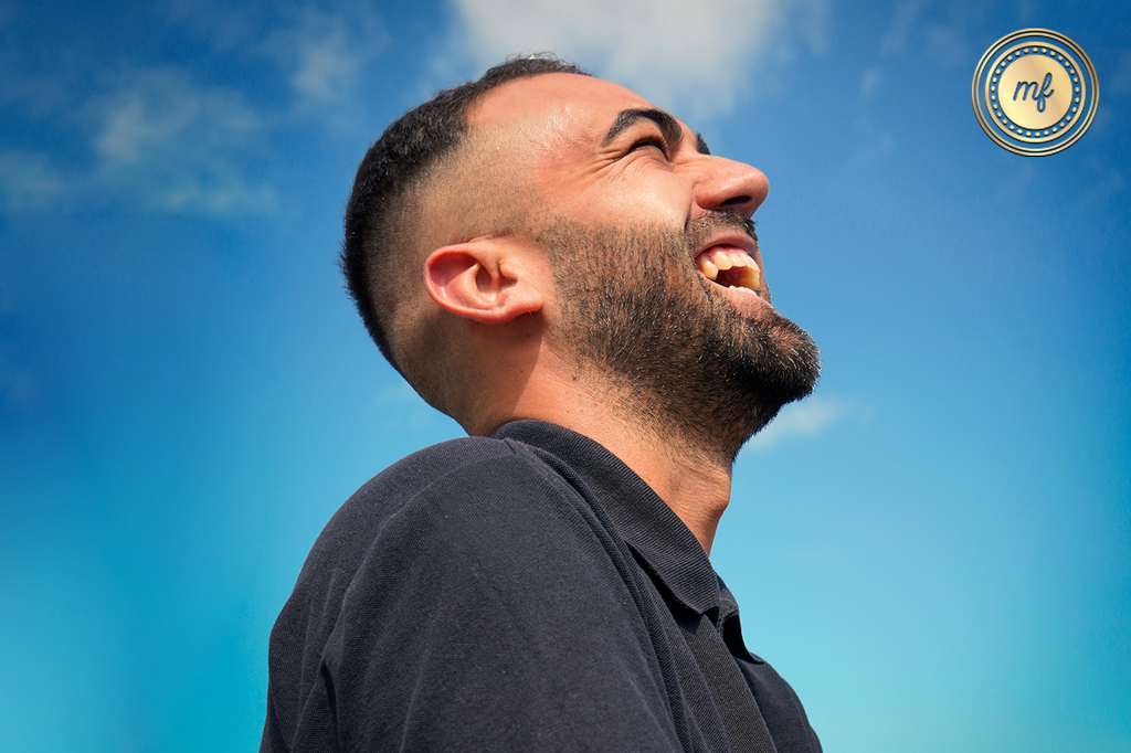 A man with a beard, wearing a dark shirt, laughs joyfully against a bright blue sky with clouds