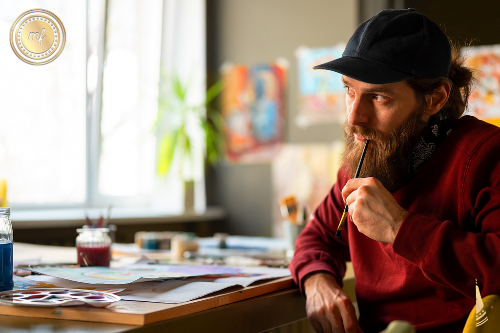 A man thinking, holding a paintbrush, with various painting supplies spread out on a table