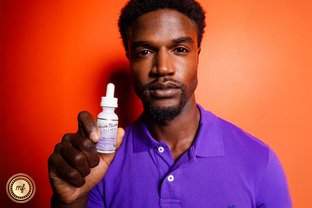 A man holding a Mellow Fellow WELLNESS: The Rest Blend tincture in his hand stands against an orange wall