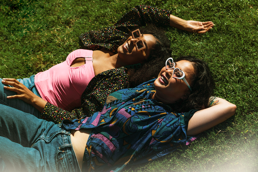Two women lie on a green field, wearing sunglasses and smiling as they enjoy themselves