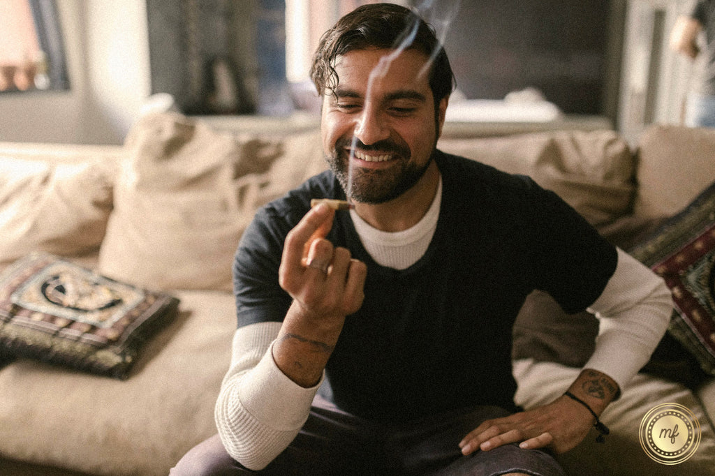 A man is holding a cannabis joint in his hand and smiling in his room