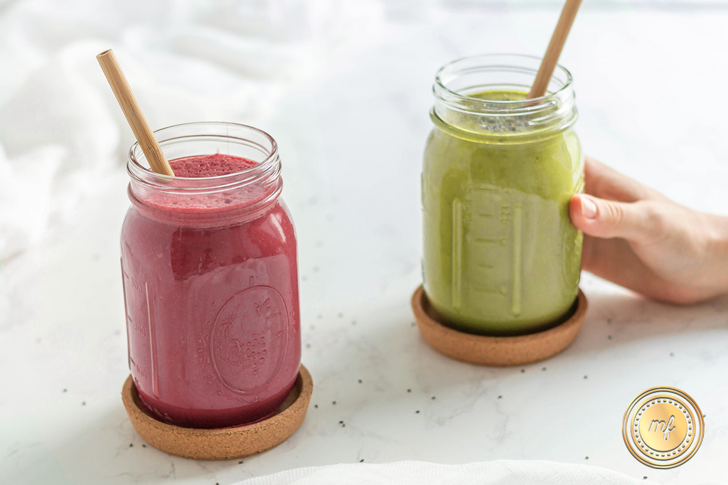 Two mason jars on a white surface contain smoothies - one red, one yellow with bamboo straws in each, as a hand holds the yellow smoothie jar