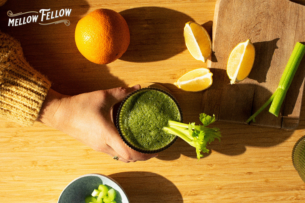 A hand holds a green smoothie with celery in a glass on a wooden surface. Beside it are sliced lemons, an orange, and celery stalks, conveying freshness.