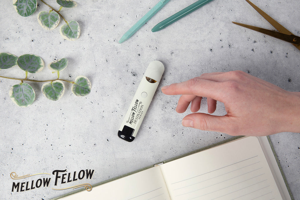 A Mellow Fellow DESTINATION vape device rests beside a notebook, a sprig, and a pen on a gray surface