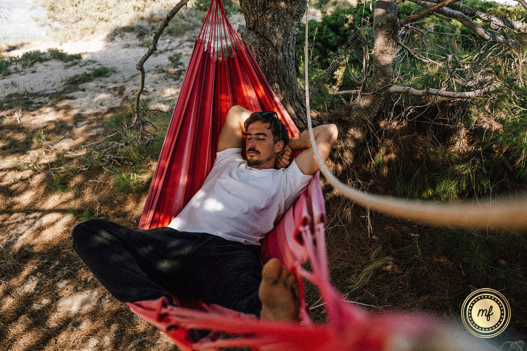 A man relaxes in a red hammock beneath the trees, eyes closed in serene ease as dappled sunlight filters through the leaves