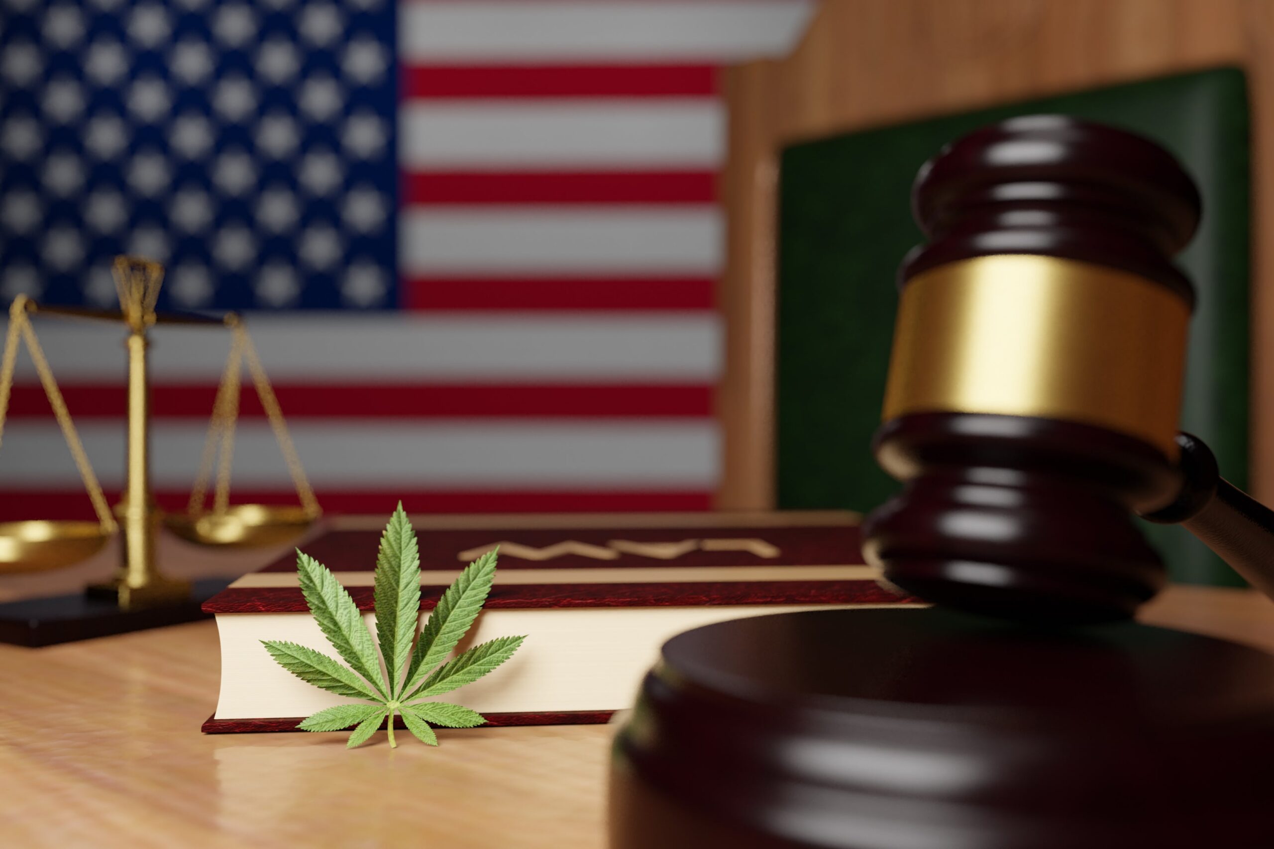 A gavel and a law book sit on a table with a cannabis leaf on the surface. In the background, there is a blurred American flag and a set of scales, symbolizing the intersection of law and cannabis legislation.