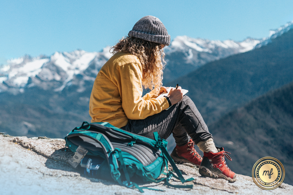 A lady sits on a mountain peak with snowy mountains in front of her.
