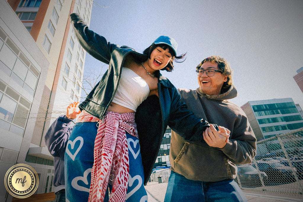 A joyful outdoor scene shows two smiling people-one in a leather jacket and cap playfully jumping, the other in a hoodie holding their hand-set against urban buildings and a clear sky