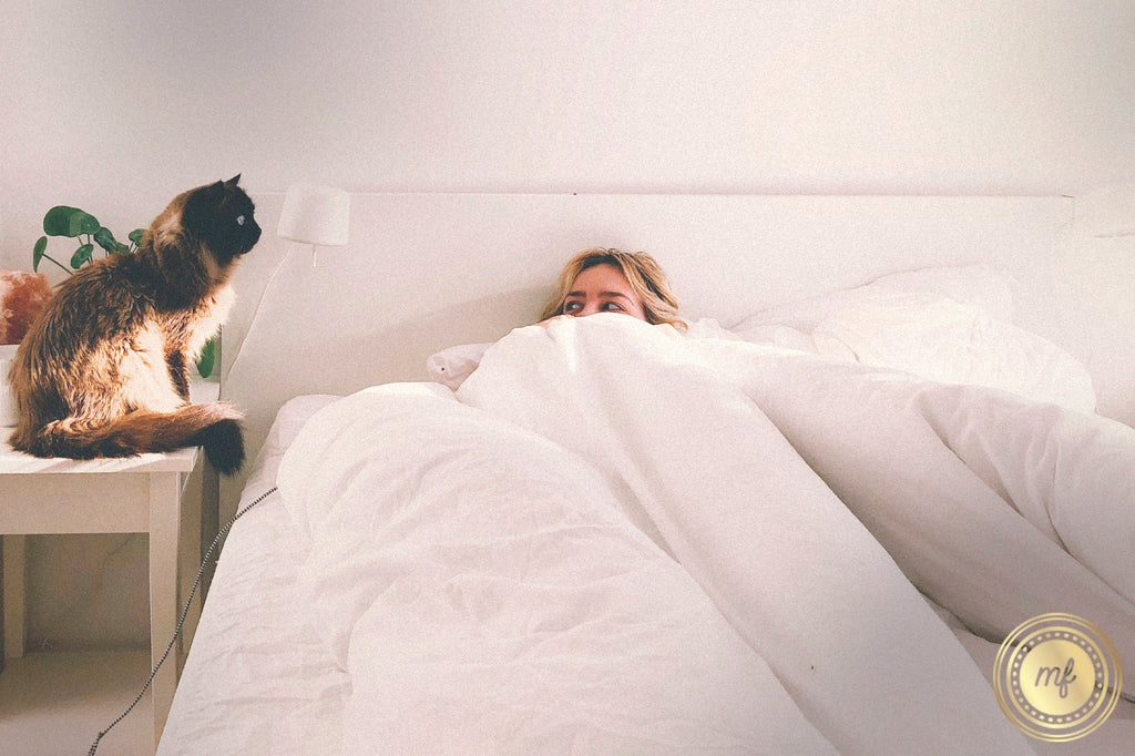 A lady wakes up from sleep, lying in her bed, covered with a white comforter, while a pet cat sits beside her on a table