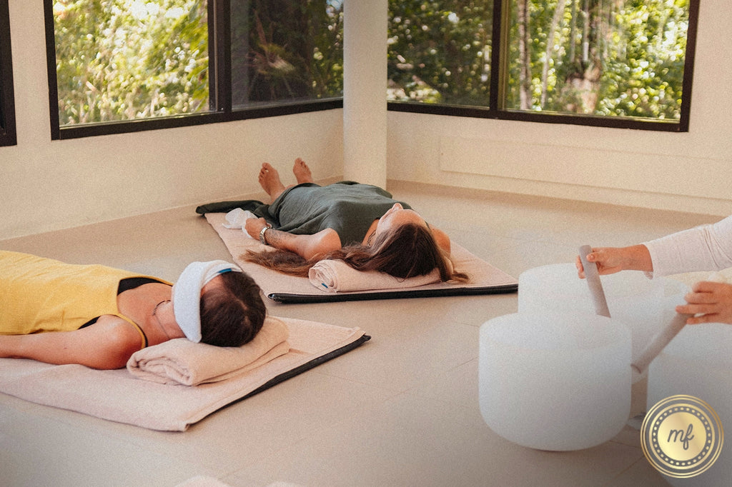 Two people lie on mats, meditating with eye covers on, deeply relaxed, while another person beside them plays white singing bowls