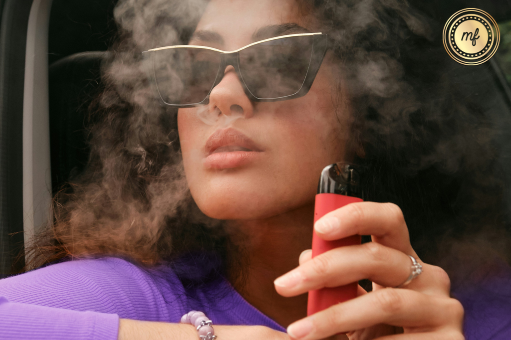 A lady wearing sunglasses, holding a vape cartridge in her hand, and exhaling smoke